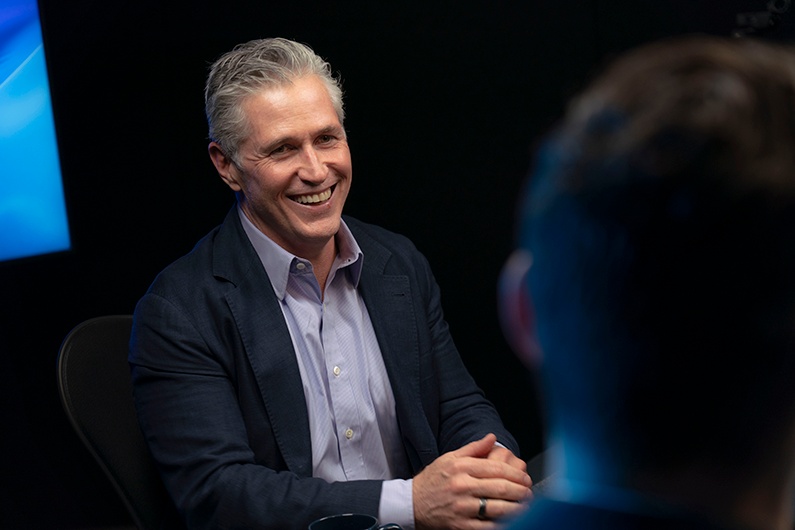 Mike Kelly speaks during a podcast recording in a studio setting, seated across from another participant.