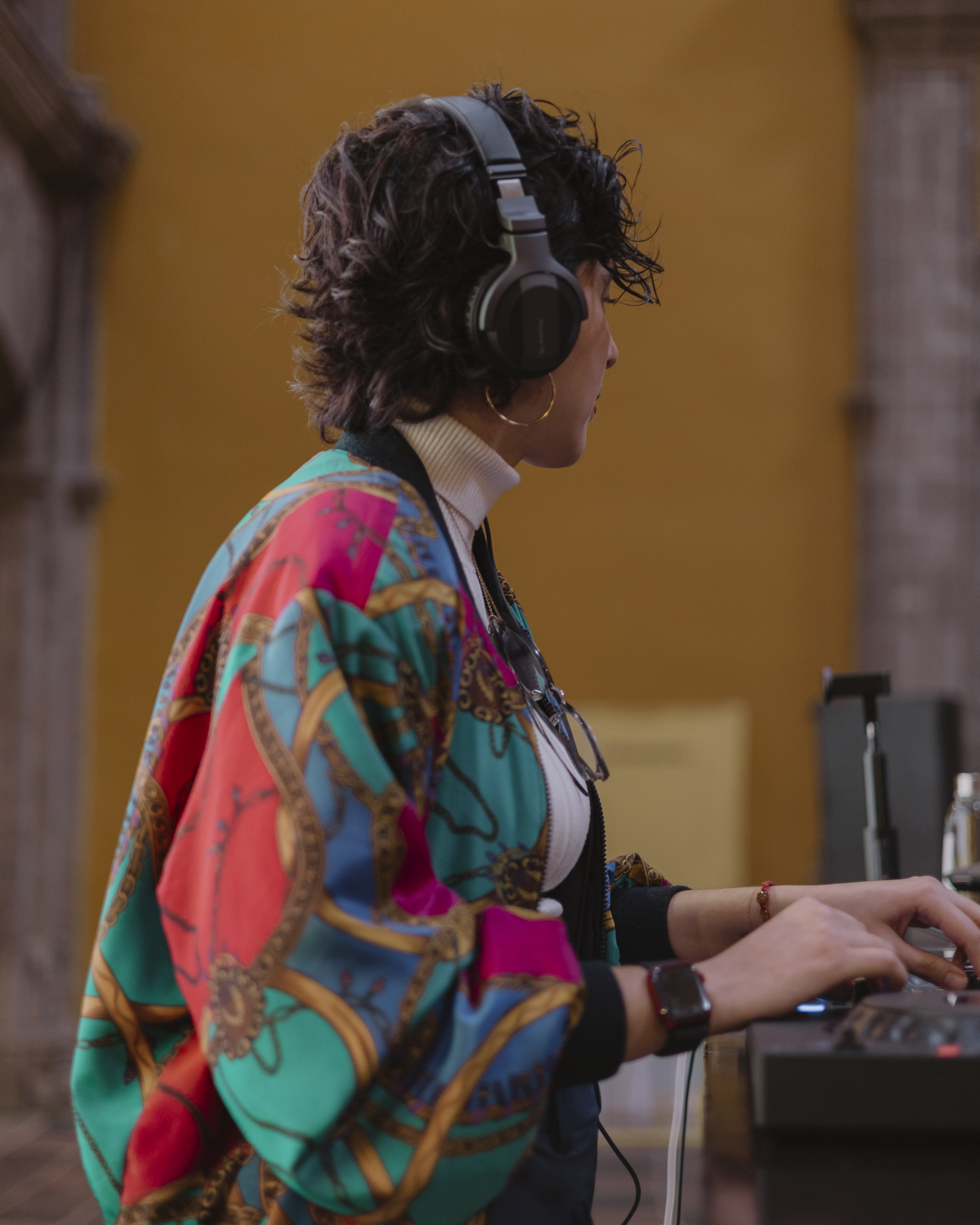 Person with curly hair wearing headphones and a colorful patterned jacket, working with DJ equipment indoors.