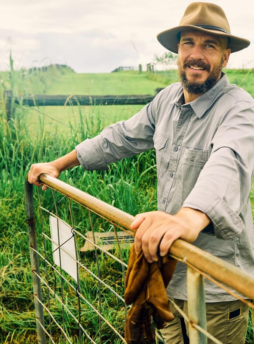 Smiling man wearing a brown hat and gray shirt leaning on a metal gate with green grass and a wooden fence in the background.
