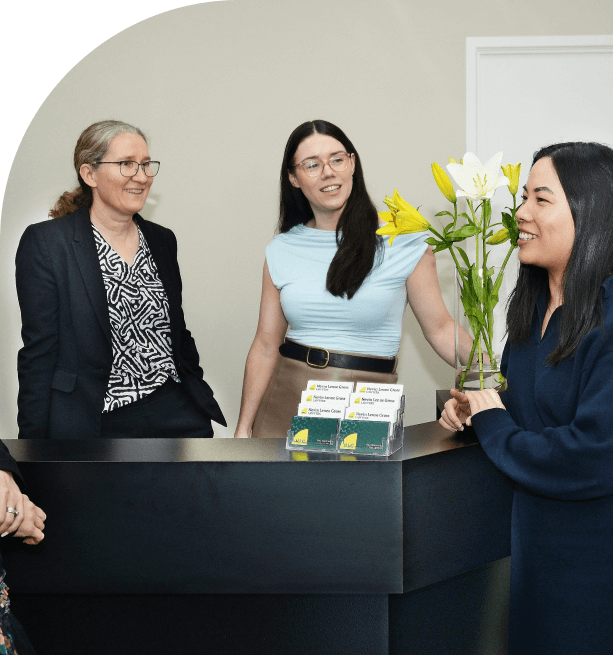 Three women smiling and talking around a black reception desk with yellow and white lilies in a vase and business cards displayed.