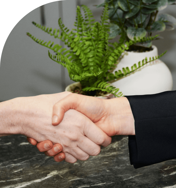 Two people shaking hands over a dark marble surface with green potted plants in the background.