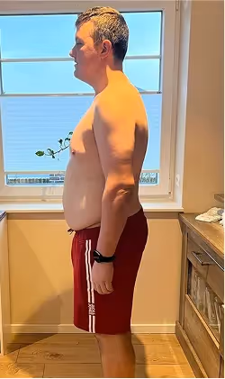 Ein Mann ohne Hemd und in roten Shorts steht seitlich vor einem Fenster.