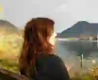 Frau mit lockigen Haaren blickt auf einen See umgeben von Bergen.