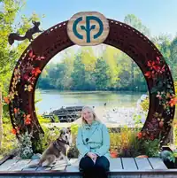Eine Frau mit Hund sitzt vor einem runden Metallbogen mit See und Bäumen im Hintergrund.