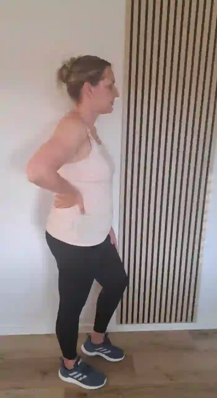 Eine Frau mit hochgesteckten Haaren und in Sportkleidung steht seitlich und hält eine Hand am Rücken.