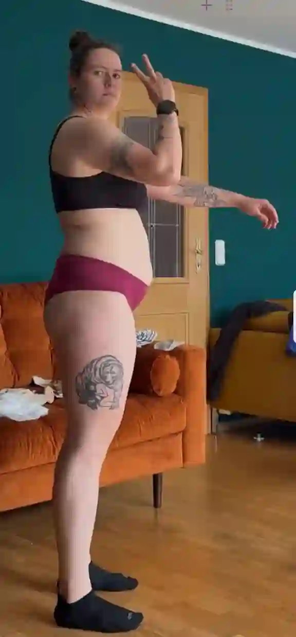 Eine Frau in Sport-BH und Unterwäsche steht seitlich mit ausgestreckten Armen vor einem Sofa.