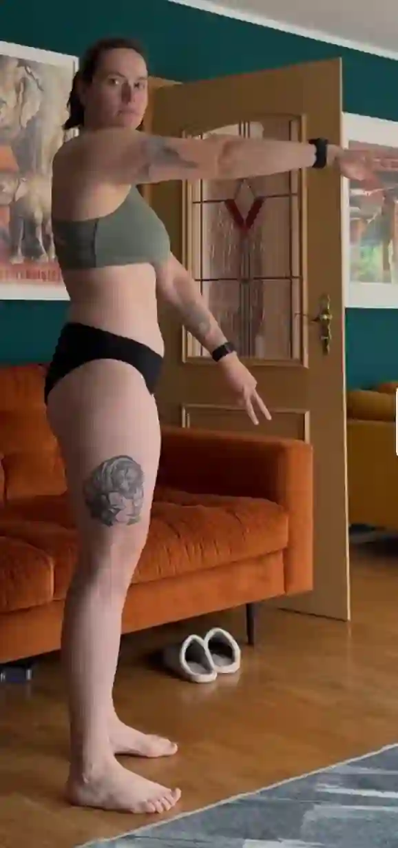 Eine Frau in Sport-BH und Unterwäsche steht seitlich mit ausgestreckten Armen vor einem Sofa.