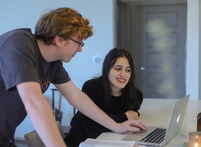 A man and a woman sitting side-by-side at a wooden table in a bright, modern workspace, looking together at a laptop screen while engaged in a collaborative discussion.