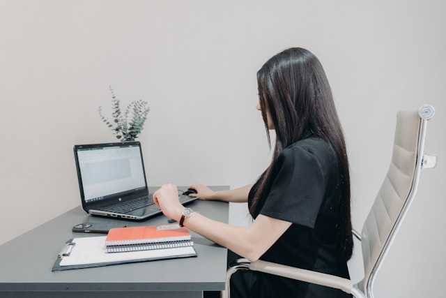 A woman sitting at a desk and focusing on her laptop screen, with open notebooks and a pen nearby in a bright, organized home office setting.