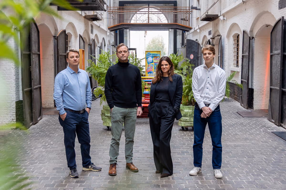 Vier mensen staan in een rij in een oude industriële gang met bakstenen muren en planten op de achtergrond.