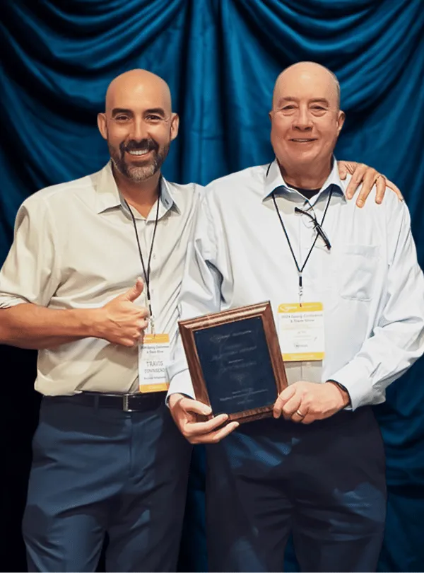 Two men standing in front of a blue curtain, one holding a plaque award and the other giving a thumbs-up.