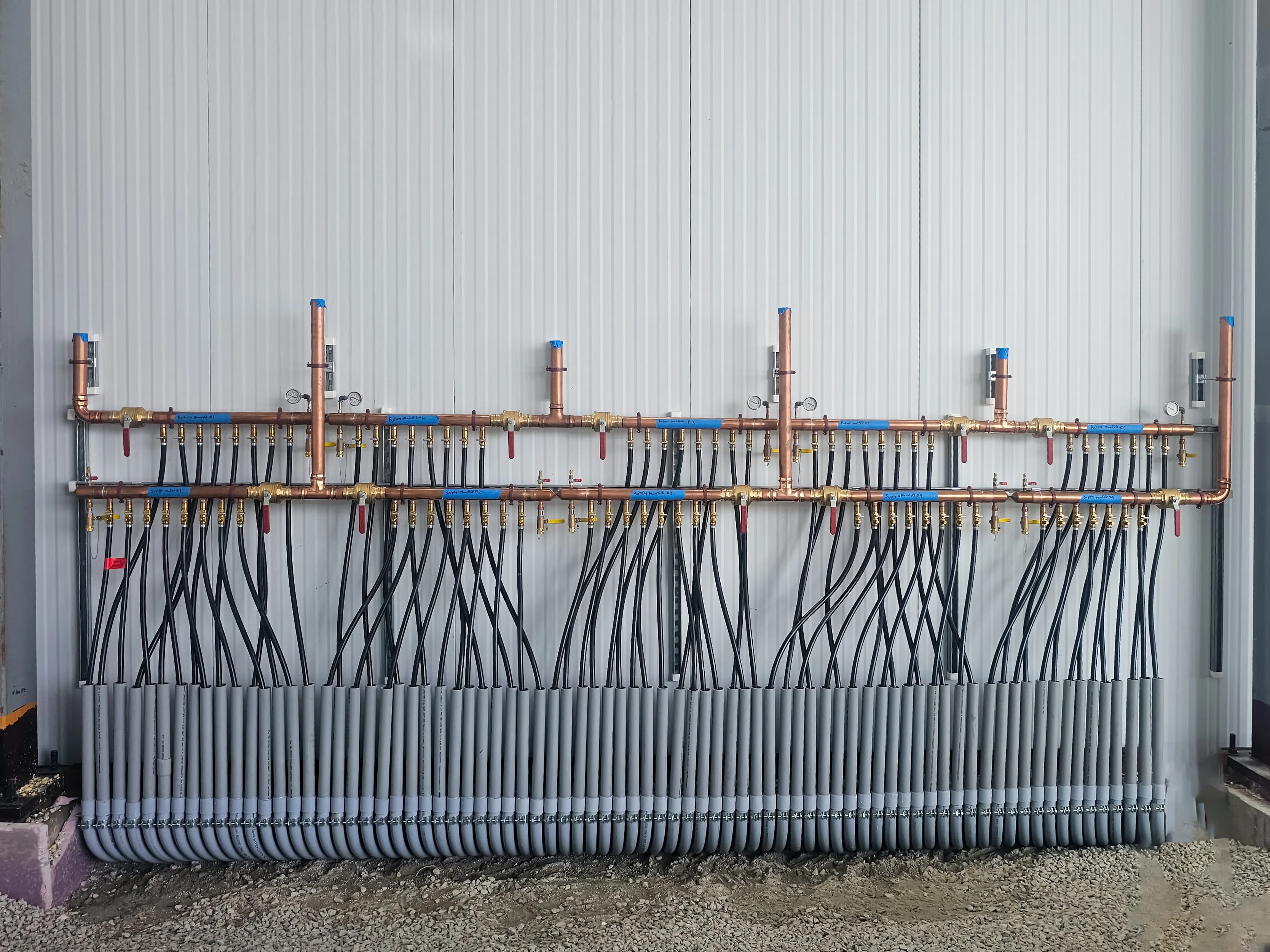 Copper plumbing manifold with multiple black tubes and gray protective covers installed on a white wall in a construction setting.