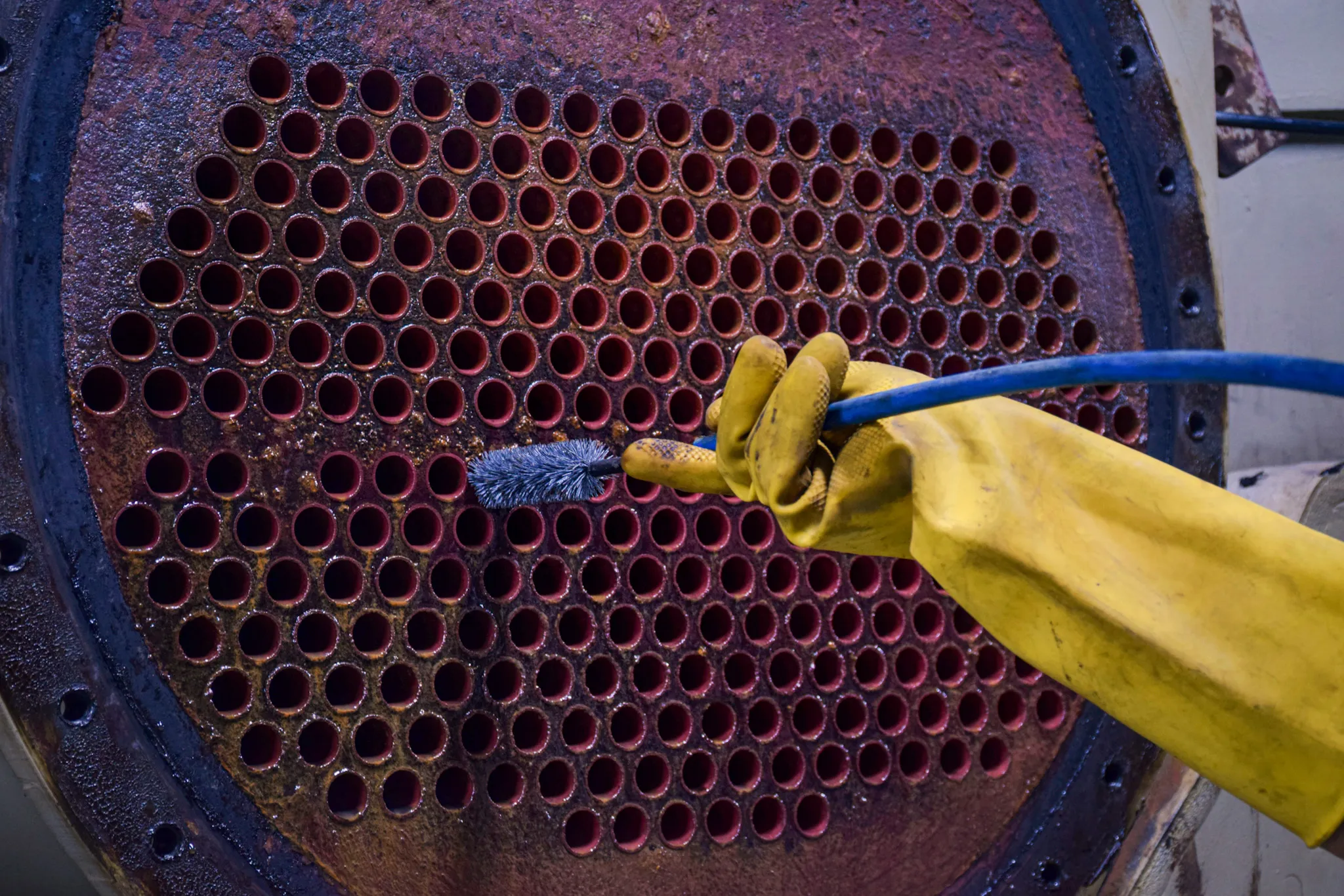 Hand wearing a yellow rubber glove cleaning rusted metal tubes with a blue brush.