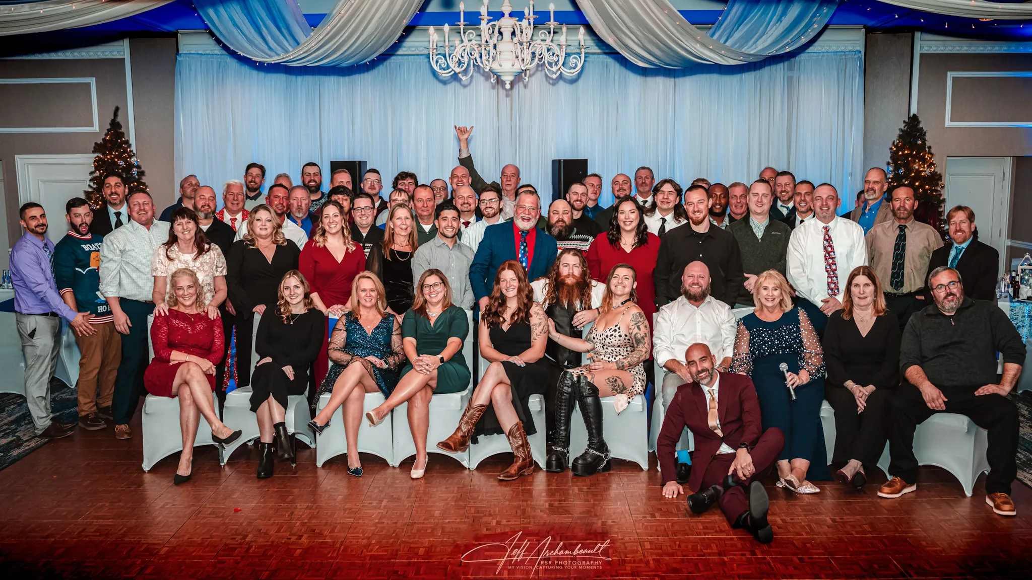Large group of men and women dressed formally posing together in a decorated room with blue drapes and chandeliers.