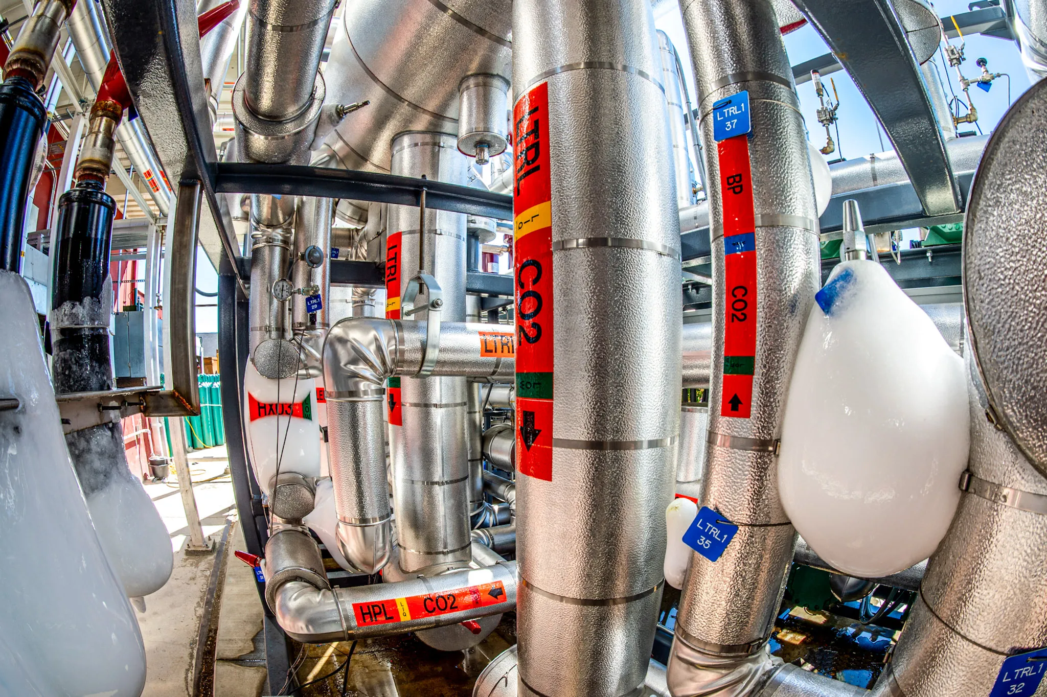 Industrial pipes and valves with bright red and blue labels indicating CO2 and other specifications, some pipes covered in ice.