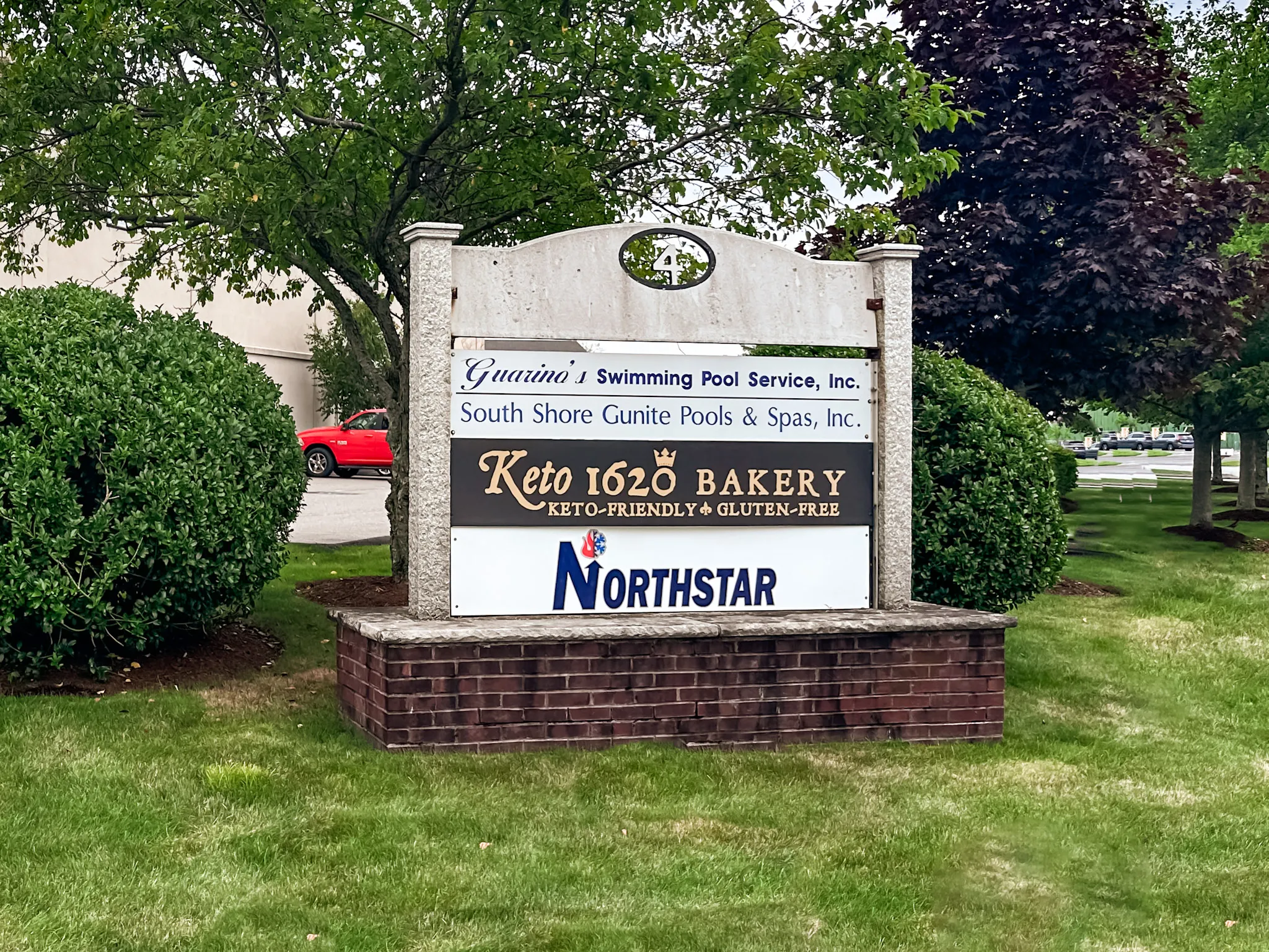 Stone and brick signboard with business names including Guarino's Swimming Pool Service, South Shore Gunite Pools & Spas, Keto 1620 Bakery keto-friendly gluten-free, and Northstar, surrounded by greenery and trees.