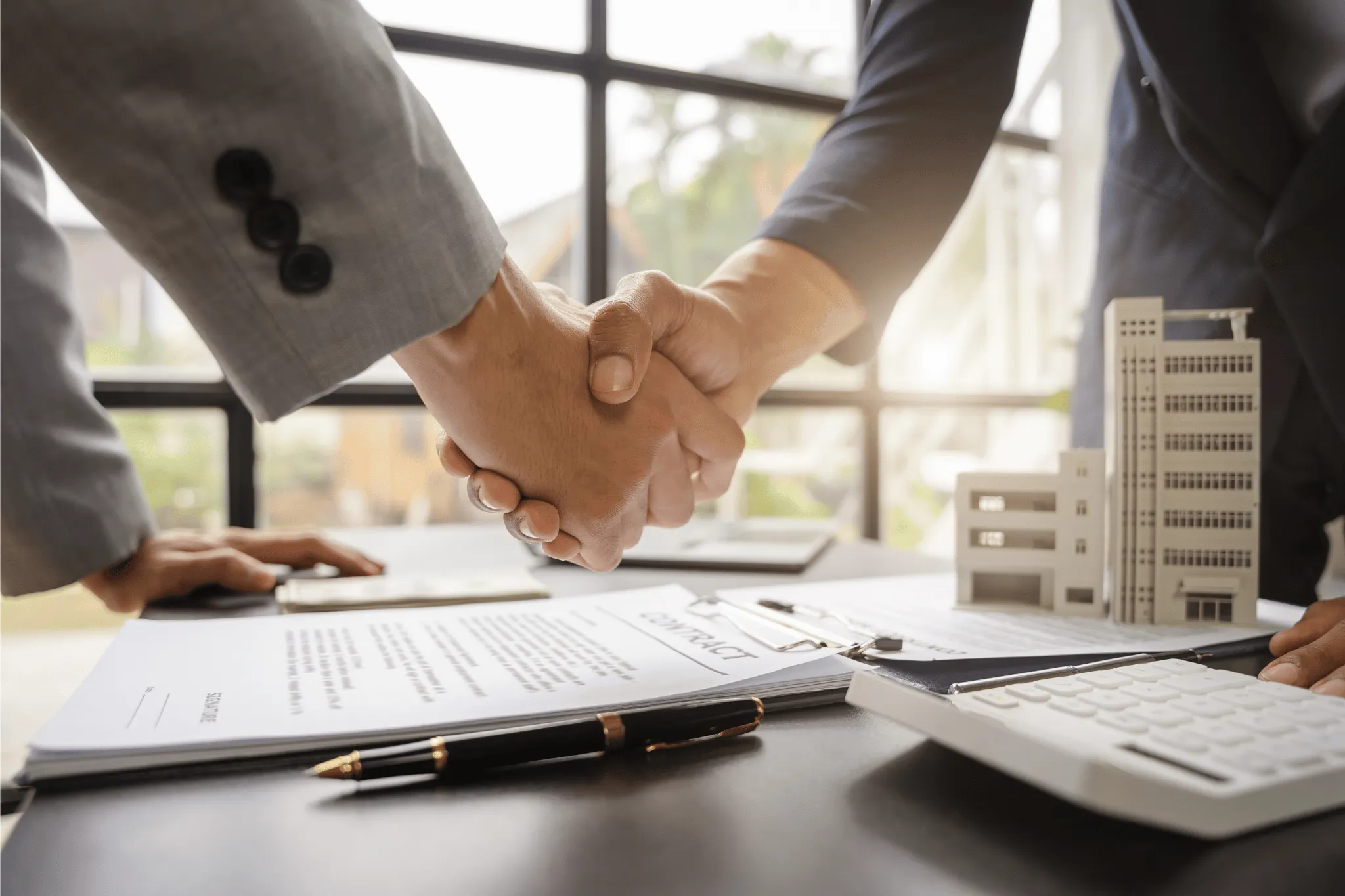 Two people shaking hands over a contract on a desk with a pen, calculator, and miniature building models.