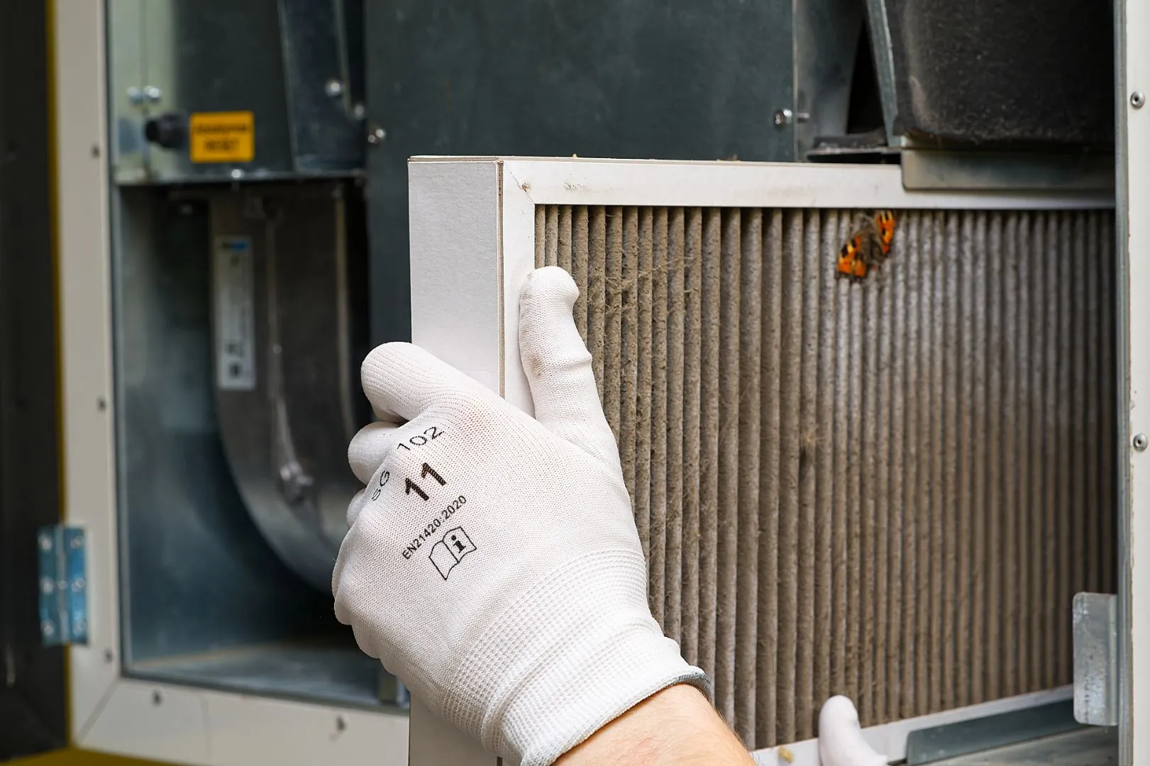 Close-up of white-gloved hands removing a dirty pleated air filter from an HVAC unit with a small butterfly resting on it.