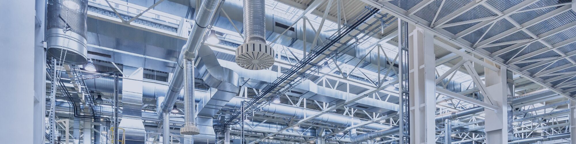 Silver metal air ducts and pipes installed on a high ceiling inside an industrial building.