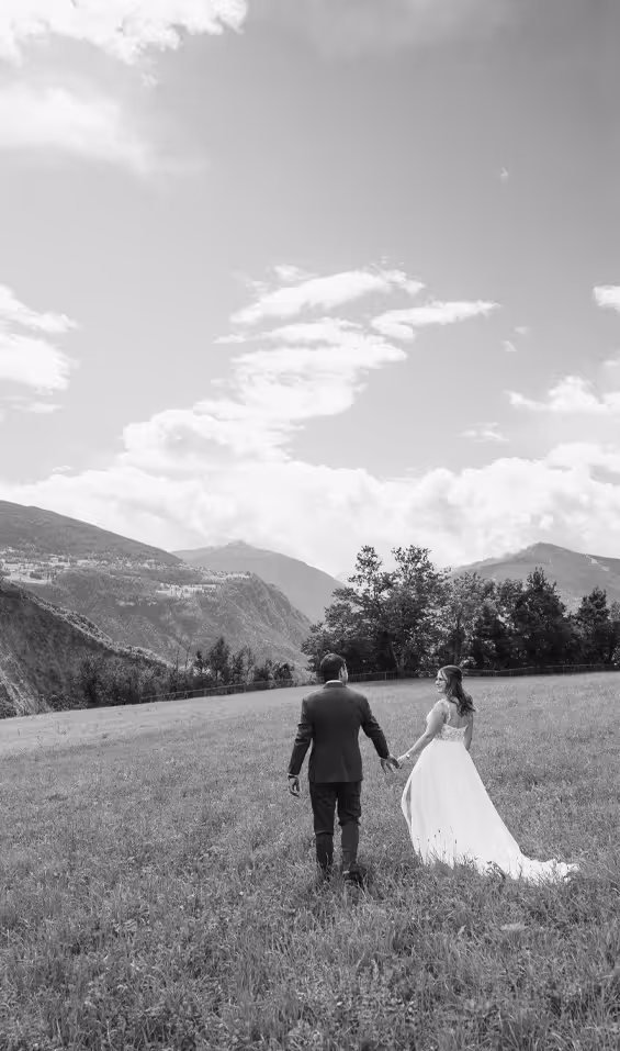 Un couple de mariés marche main dans la main dans un champ avec des montagnes en arrière-plan.