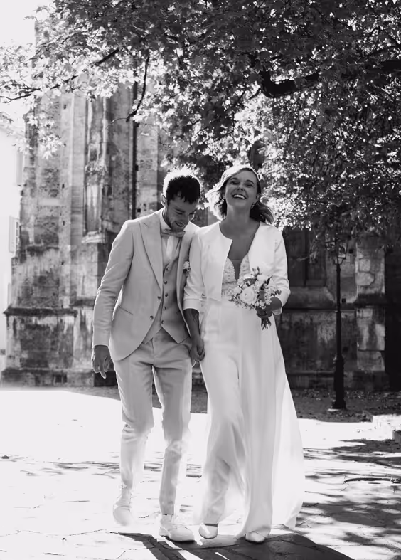 Couple de mariés souriants marchant main dans la main devant un bâtiment ancien sous des arbres.