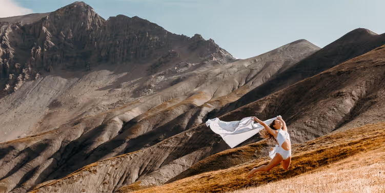 Femme en tenue blanche courant et tenant un tissu flottant sur une colline herbeuse avec des montagnes rocheuses en arrière-plan.