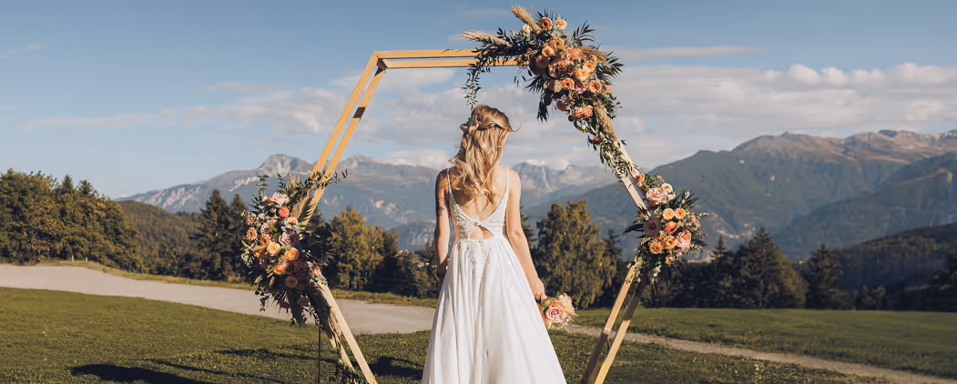 Femme en robe blanche de mariée de dos tenant un bouquet devant une arche hexagonale fleurie dans un paysage montagneux ensoleillé.