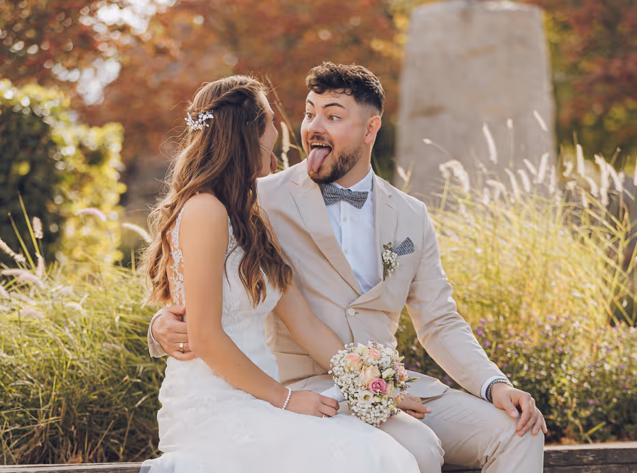 Couple de mariés assis, le marié tire la langue en regardant la mariée qui sourit.