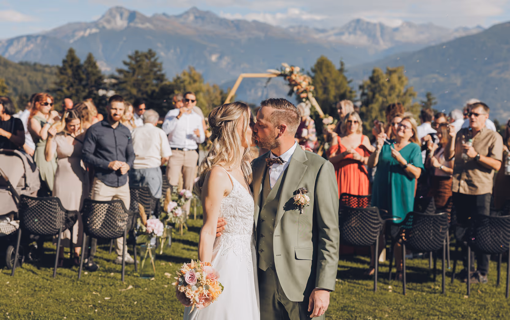 Un couple de mariés s'embrassant lors d'une cérémonie en plein air avec des invités en arrière-plan et des montagnes en arrière-plan.