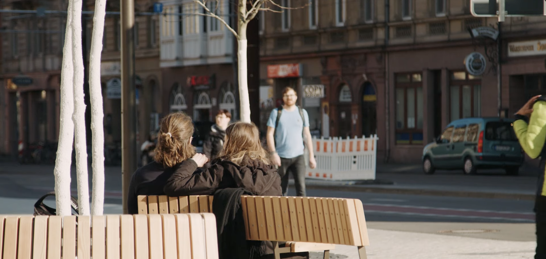 Zwei Personen sitzen auf einer modernen Holzbank in der hellen Karlsruher Innenstadt, die zukünftige Stadtentwicklung widerspiegelnd.