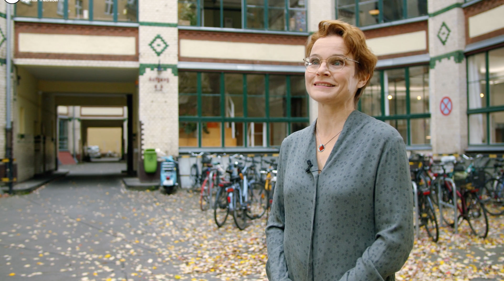 Authentischer Videostill einer lächelnden Bauingenieurin mit Mikrofon in einem urbanen Innenhof mit Altbaufassade, Fahrrädern und Herbstlaub, aus dem Recruiting-Video der Ingenieur Gruppe Bauen.