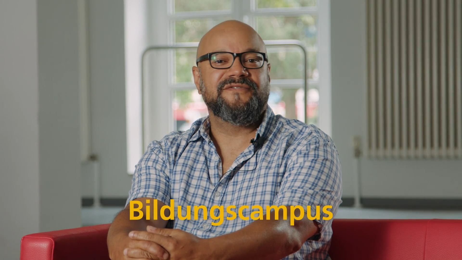 Bärtiger Mann mit Brille auf rotem Sofa, gelber Text Bildungscampus. Filmszene zu Karlsruhes Sozial- und Bildungsprojekten.