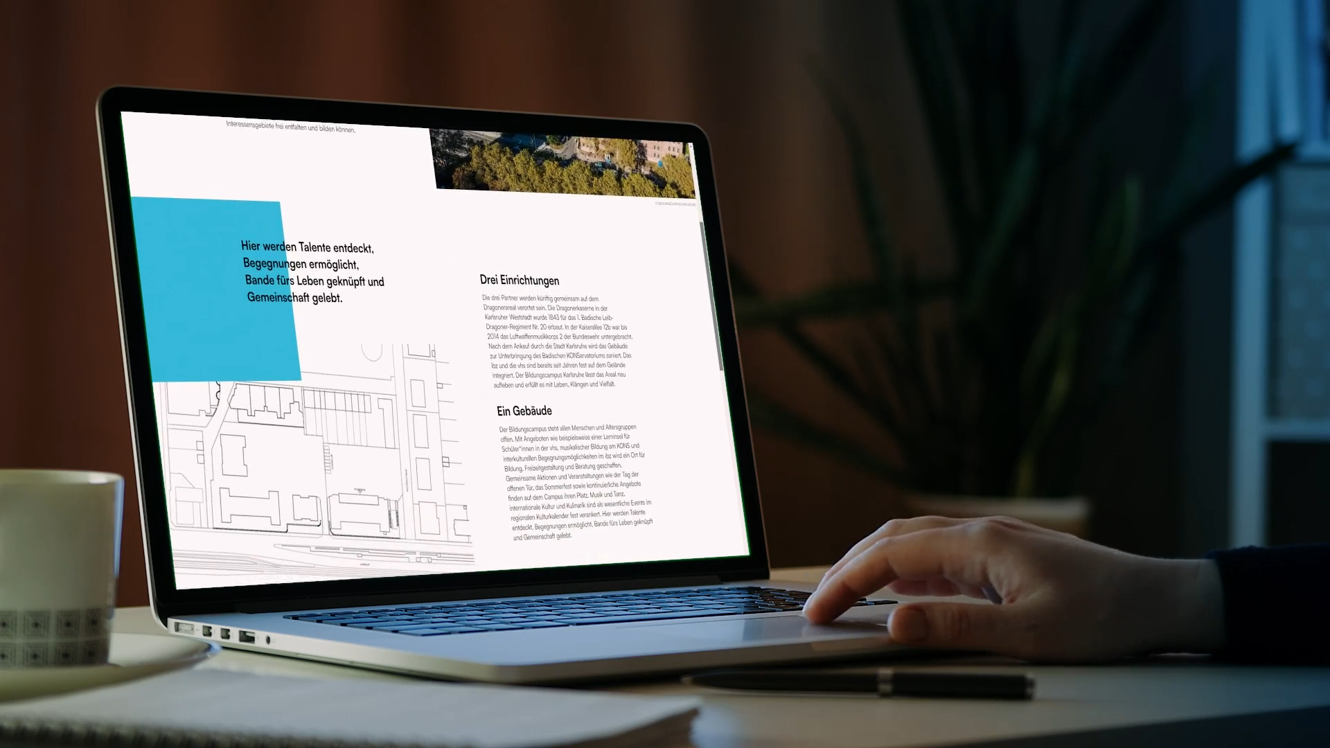 Laptop zeigt Präsentation zu Karlsruher Sozialprojekten mit Text und Planzeichnung. Eine Hand bedient das Gerät, daneben Tasse und Stift. Professionelle Ästhetik.