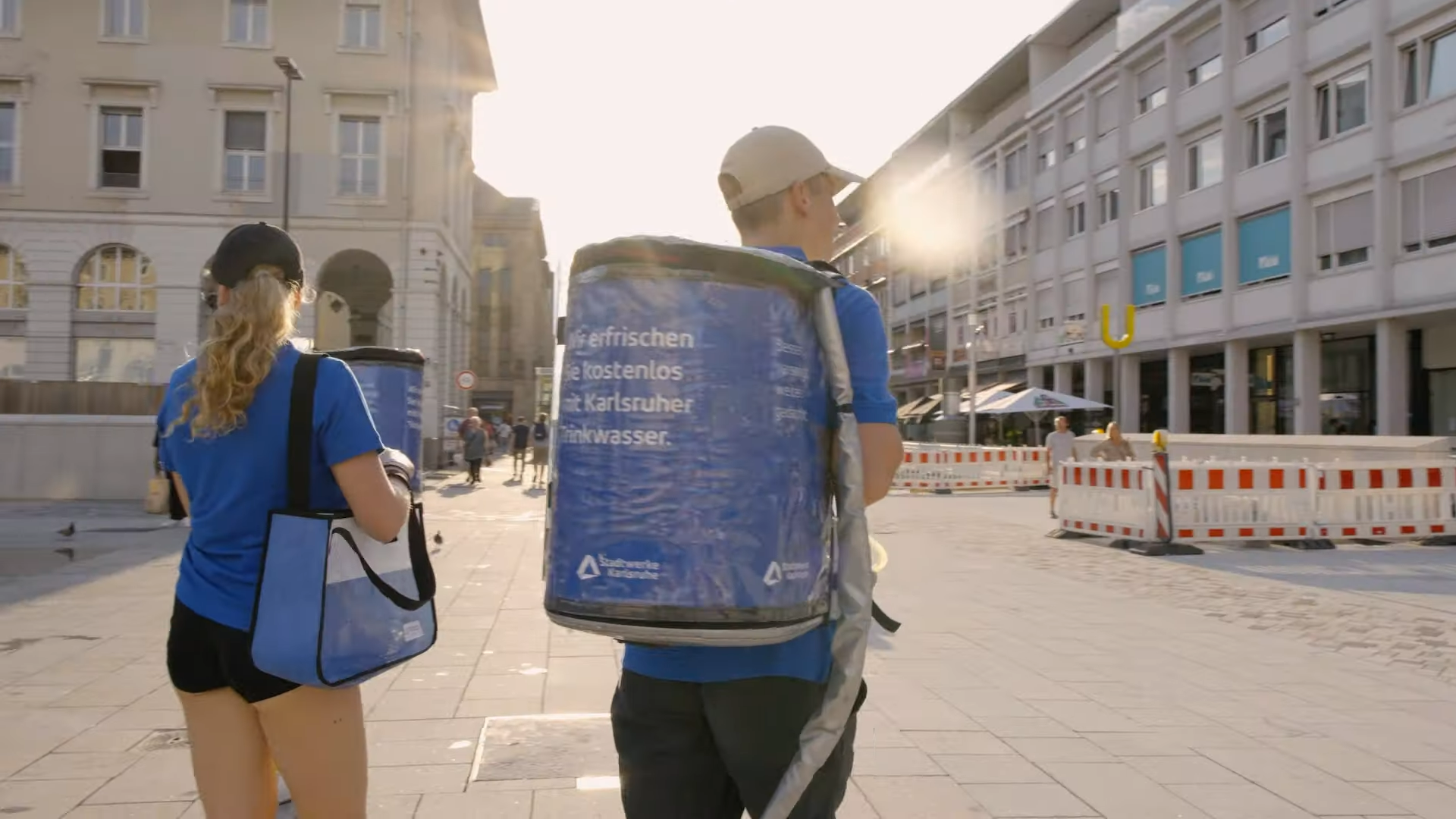 Zwei Personen bei der Trinkwasser-Promotion der Stadtwerke Karlsruhe, mit Rucksack und Tasche, auf einem sonnigen Stadtplatz.