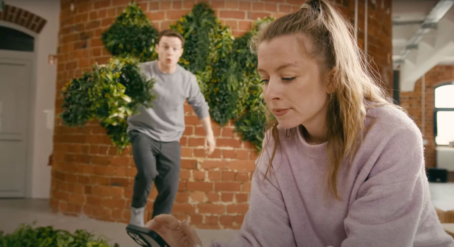 Dynamische Szene der Webserie über grüne 3D-Fassaden: Eine Frau blickt auf ihr Smartphone, während ein Mann im Hintergrund schwungvoll begrünbare Elemente vor einer Ziegelwand mit Pflanzeninstallationen trägt.