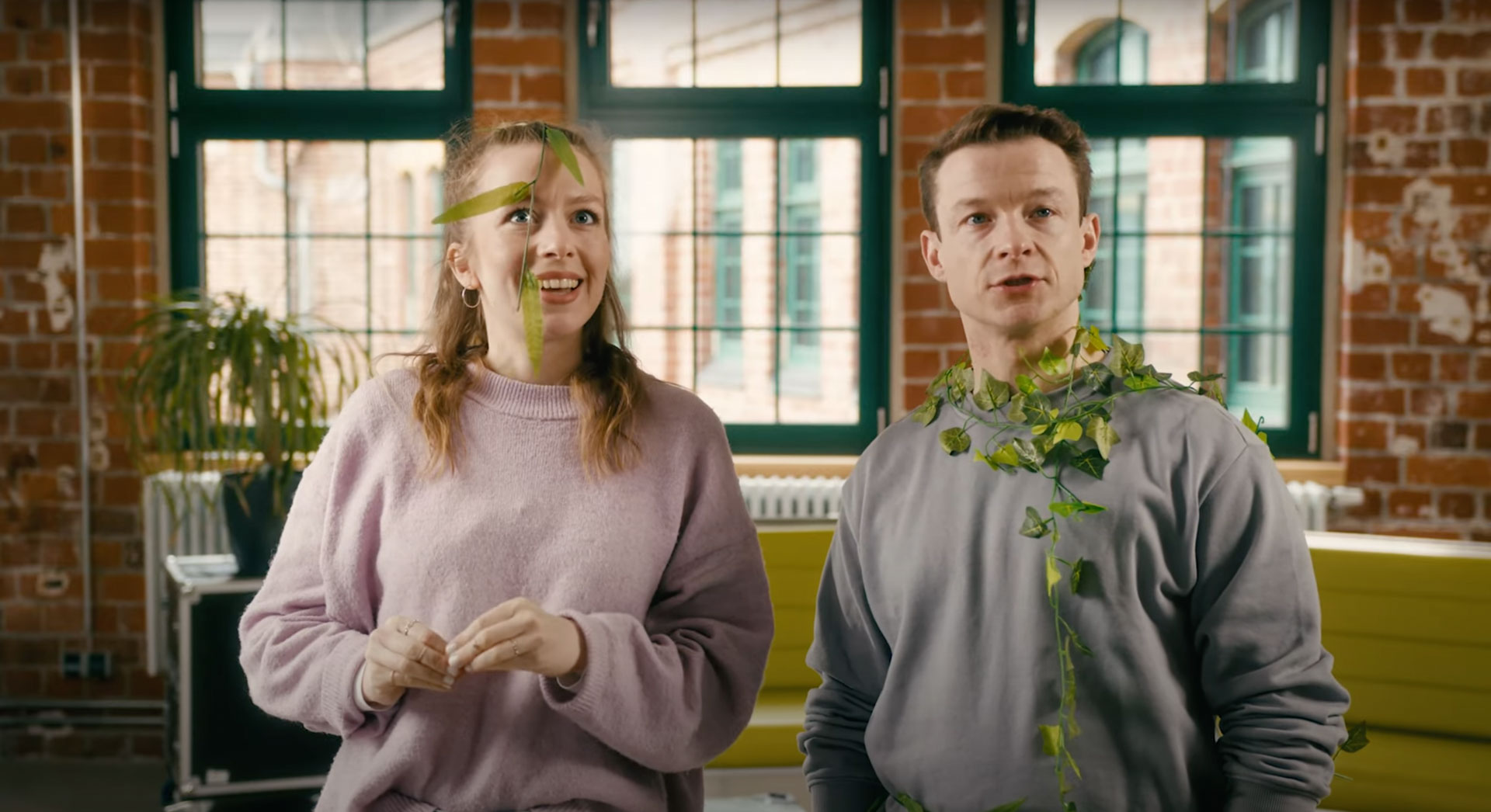 Zwei junge Menschen, mit Blättern im Gesicht und an der Kleidung, präsentieren spielerisch Forschung zu begrünten 3D-Fassaden in einem modernen Industrielook-Studio.