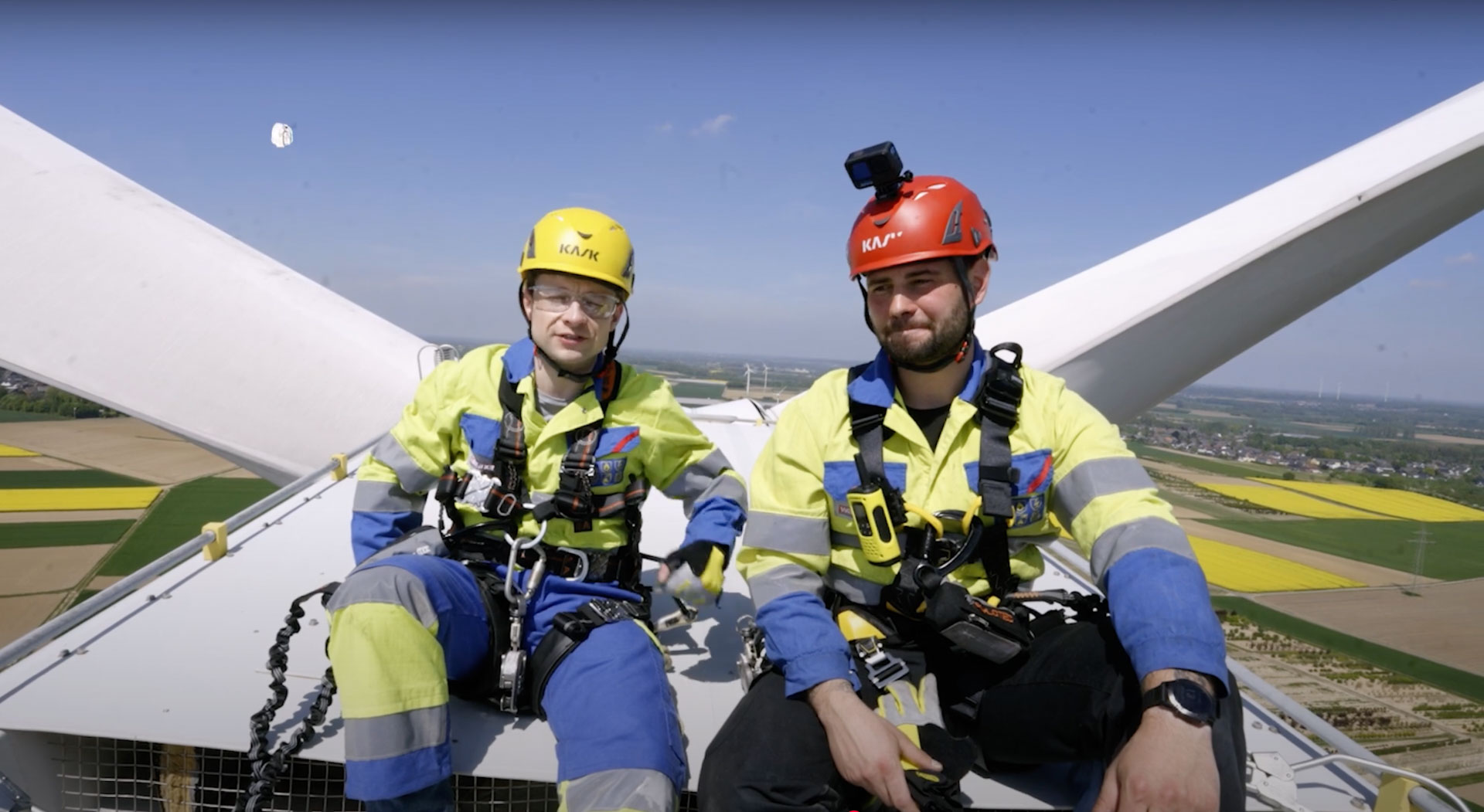 Zwei Männer in Servicetechniker-Schutzkleidung sitzen auf einer Windkraftanlage, illustrierend Berufe der Zukunft aus großer Höhe. Die Darstellung ist klar und realistisch.