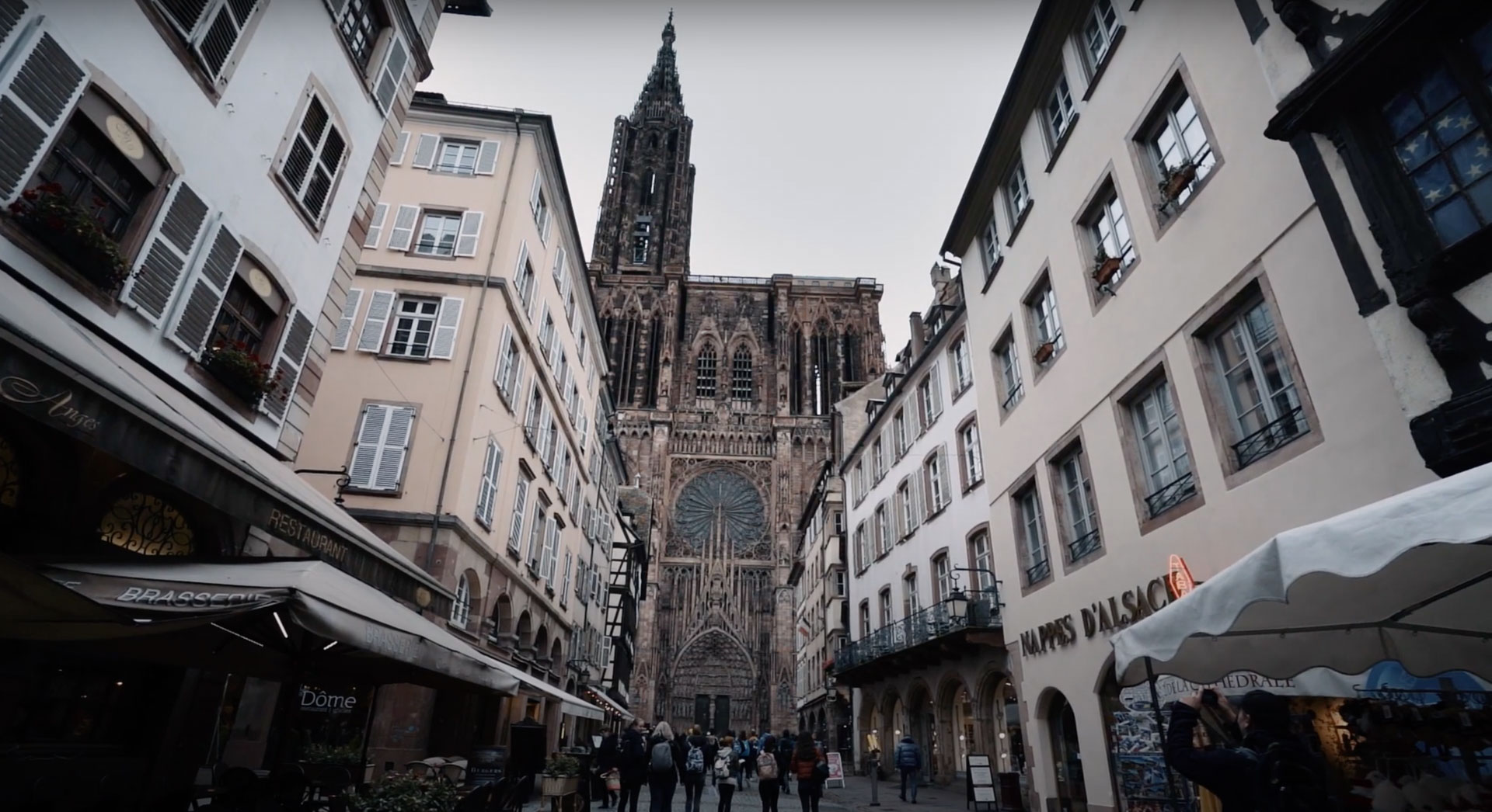Monumentale Straßburger Kathedrale am Ende einer historischen Gasse, umrahmt von Altstadtgebäuden mit Menschen, im dokumentarischen Filmstil.