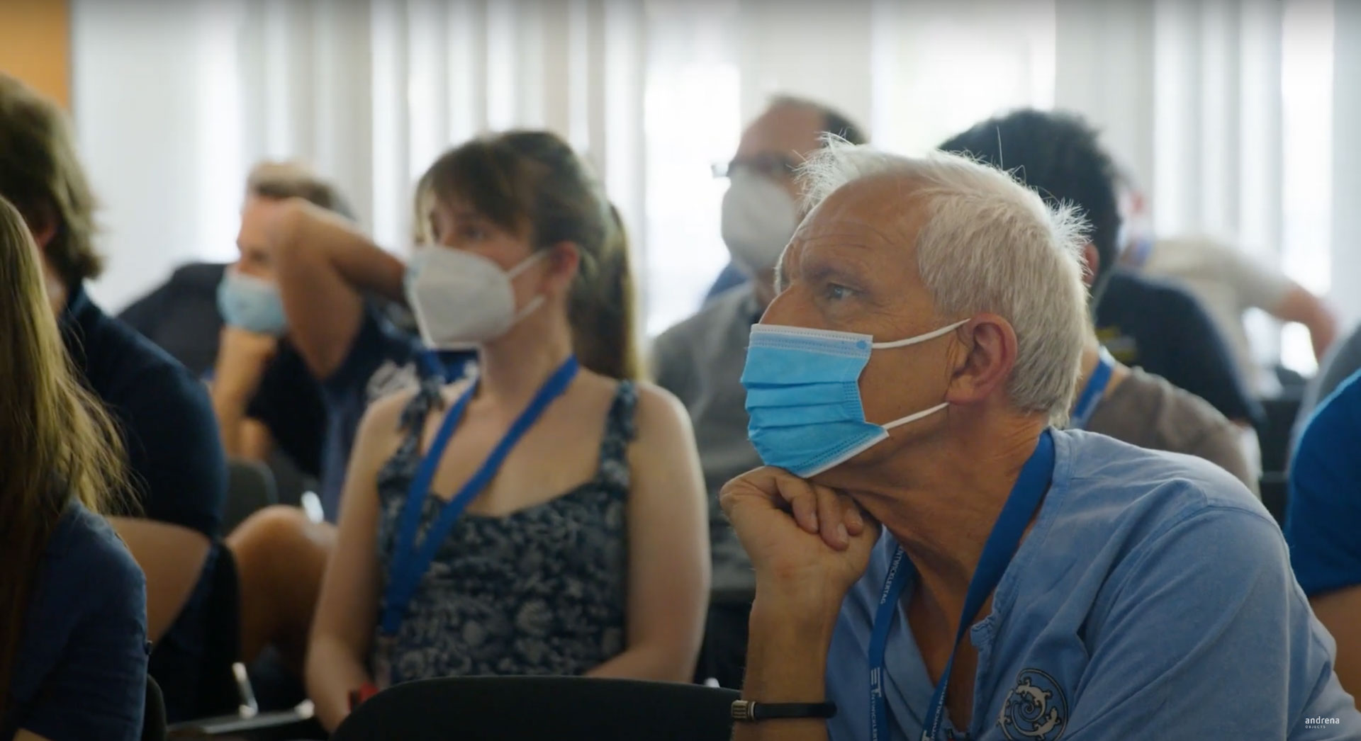 Ein älterer, maskierter Teilnehmer des Entwicklertags Karlsruhe sitzt aufmerksam in einer Konferenzreihe, flankiert von weiteren maskierten Besuchern. Das Bild fängt die konzentrierte Atmosphäre der IT-Veranstaltung ein.