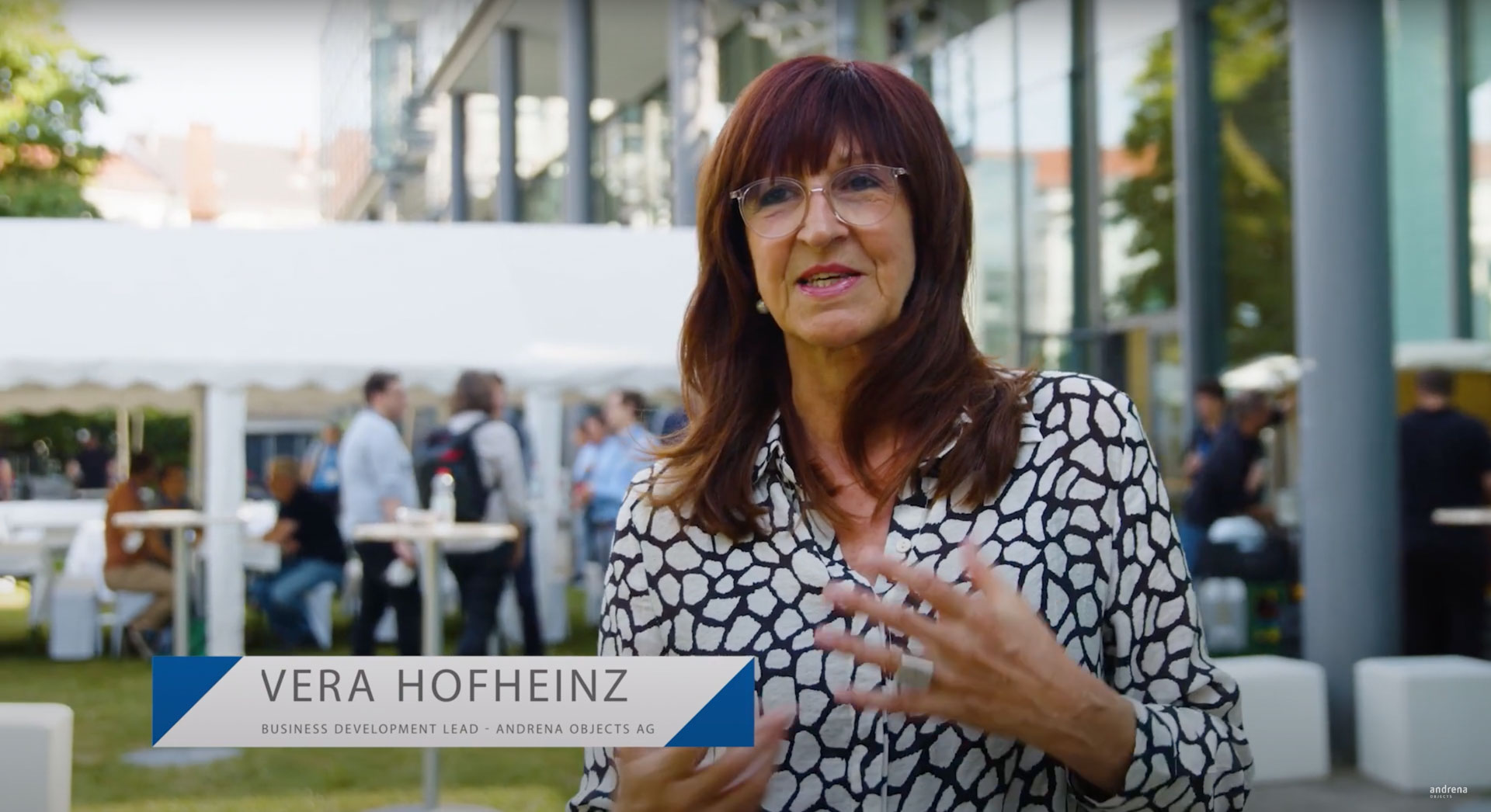 Vera Hofheinz, Business Development Lead von Andrena Objects AG, in einem professionellen Video-Statement auf dem Entwicklertag, mit lebhaftem Eventgeschehen im unscharfen Hintergrund.