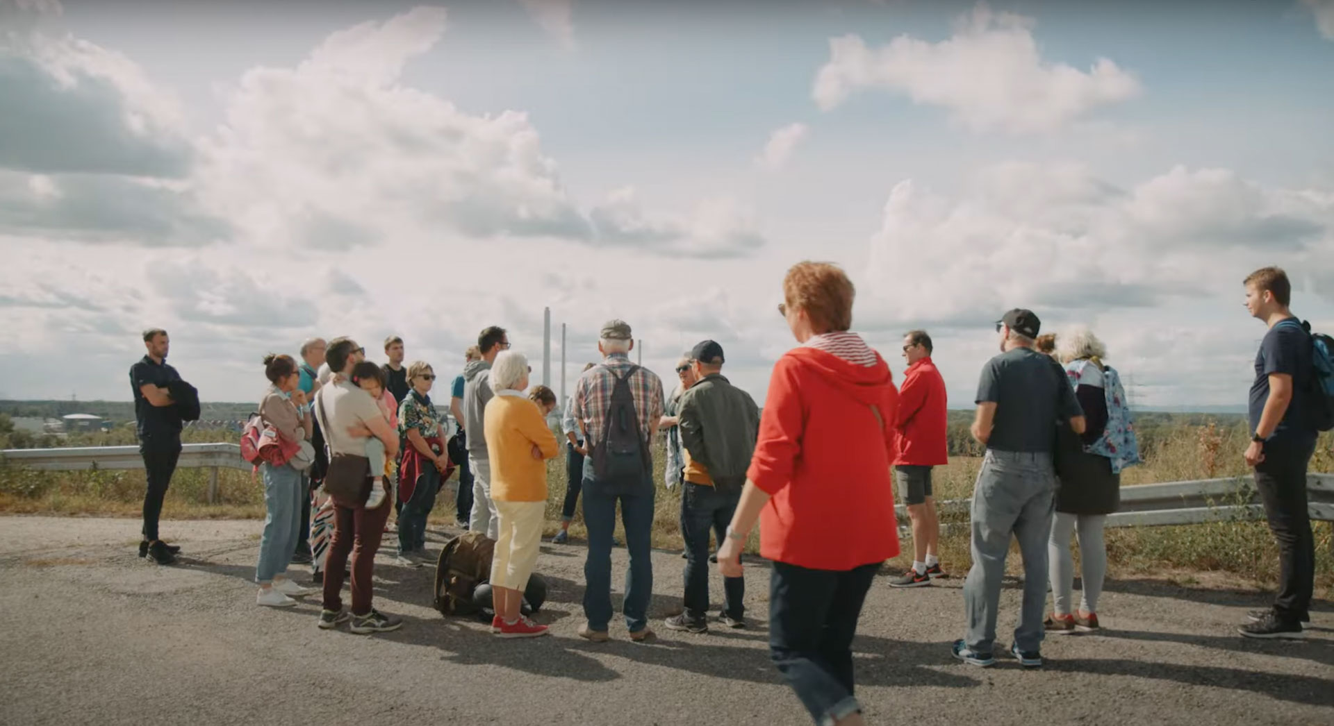 Eventfilm-Still: Eine vielfältige Gruppe steht auf dem Karlsruher Energieberg und blickt auf Industrieanlagen, dokumentierend den Energiewendetag.