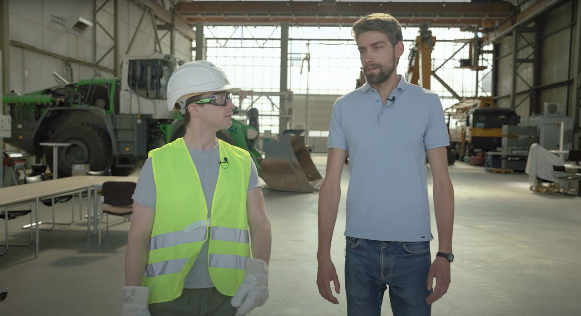Die Moderatoren Ryan und Caro in einer großen Halle mit Baumaschinen für den Erklärfilm „Baustelle der Zukunft“.