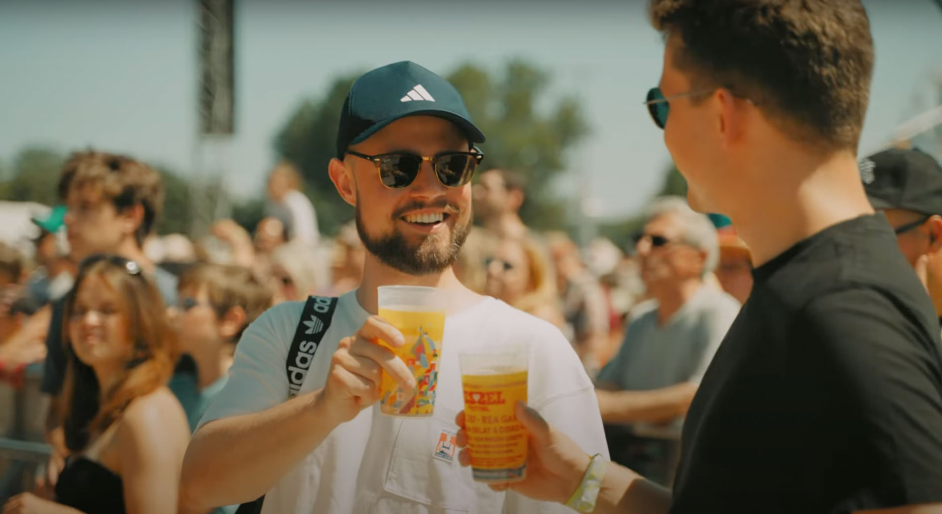 Zwei junge Männer stoßen auf dem sonnigen Kessel Festival mit Festivalbechern an, ein fröhlicher Moment in lebendiger Atmosphäre.