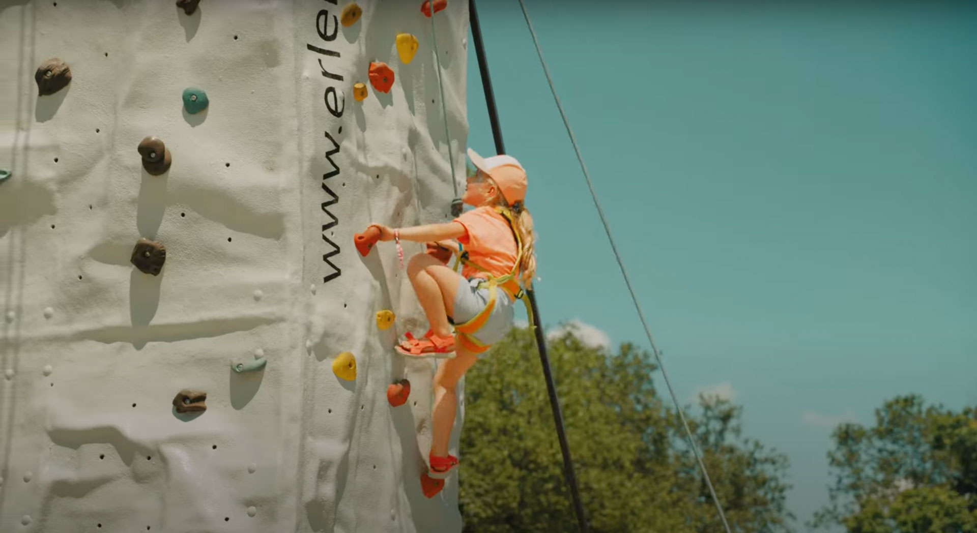 Ein Kind klettert an einer hellen Kletterwand vor blauem Himmel.