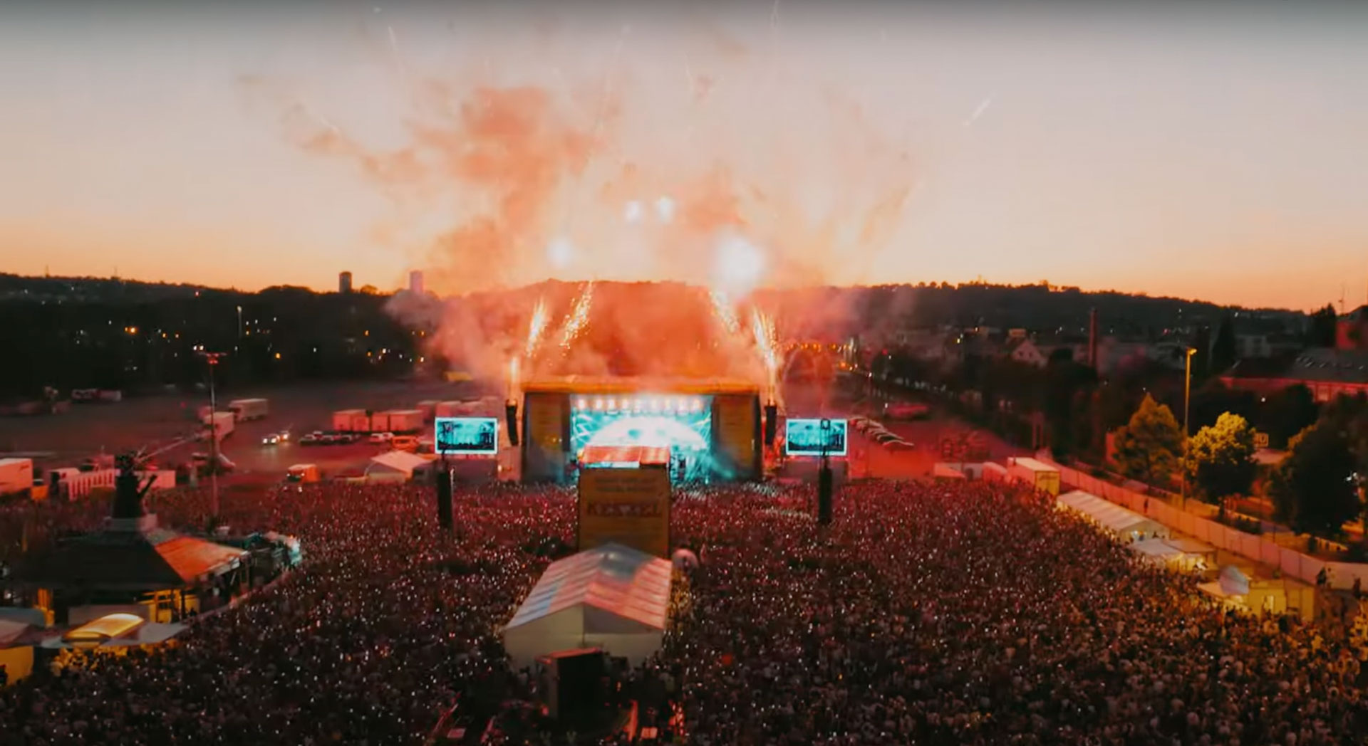 Luftaufnahme des Kessel Festivals bei Sonnenuntergang: Eine riesige Menschenmenge versammelt sich vor der beleuchteten Hauptbühne, von der Pyrotechnik aufsteigt.