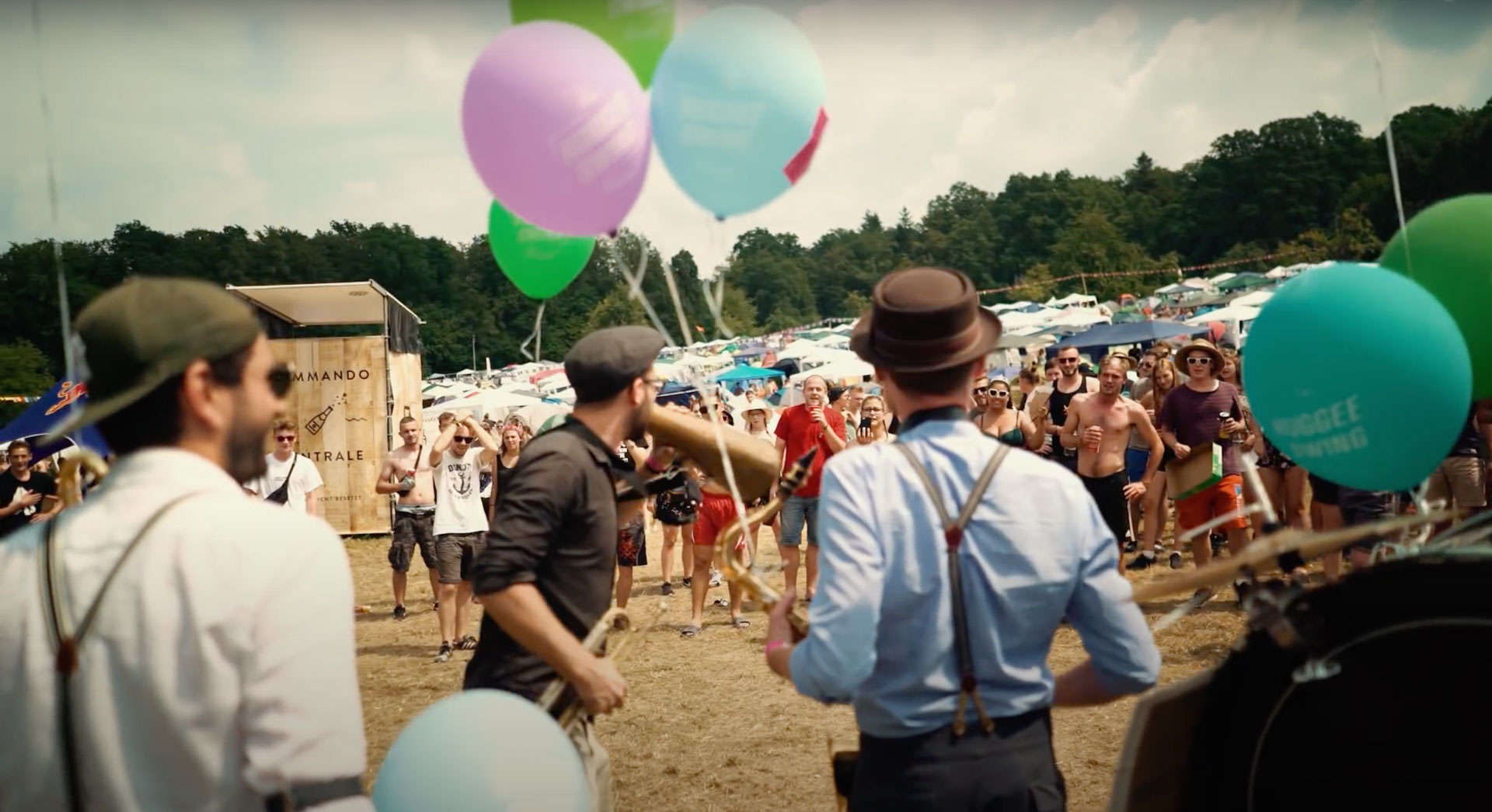Musiker spielen auf dem Happiness-Festival vor fröhlichem Publikum; bunte Ballons und Stände prägen die lebhafte Freiluftszene.