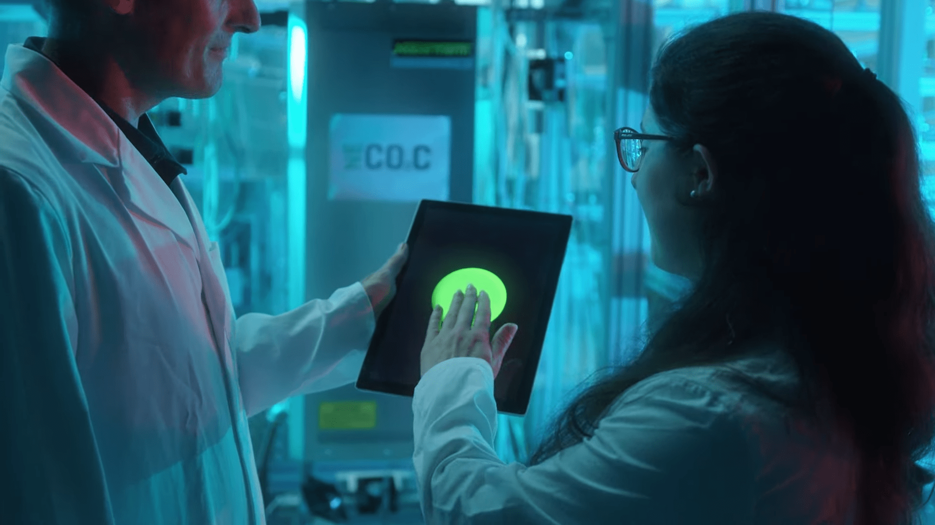 Zwei KIT-Wissenschaftler in Laborkitteln bedienen ein Tablet mit leuchtend grüner Anzeige in einem modern, blau beleuchteten Labor. Ein 'CO.C'-Schild im Hintergrund verweist auf die CO2-Technologie. Der Stil ist technisch-innovativ.