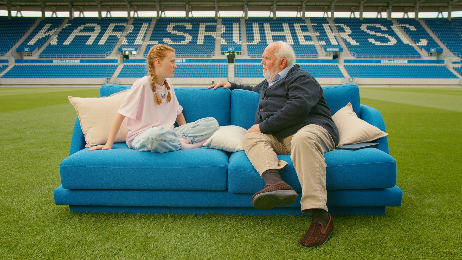 Ein Mädchen und ein älterer Mann sitzen auf einem blauen Sofa auf dem Rasen des Karlsruher SC Stadions. Eine symbolische Szene für Verbundenheit.