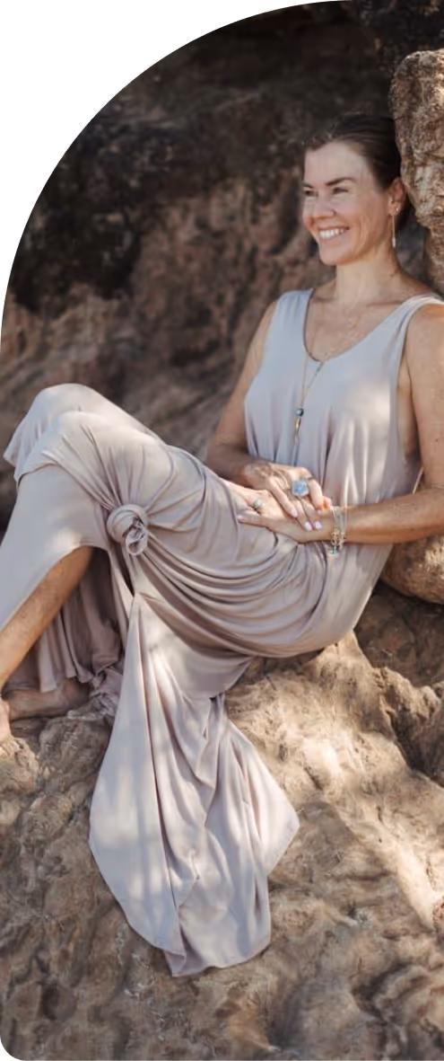 Woman in a light beige sleeveless dress sitting and smiling on rocky terrain.