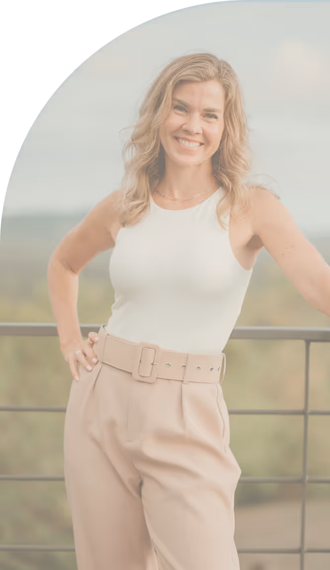 Smiling woman with wavy blond hair wearing a white sleeveless top and beige high-waisted pants standing outdoors.